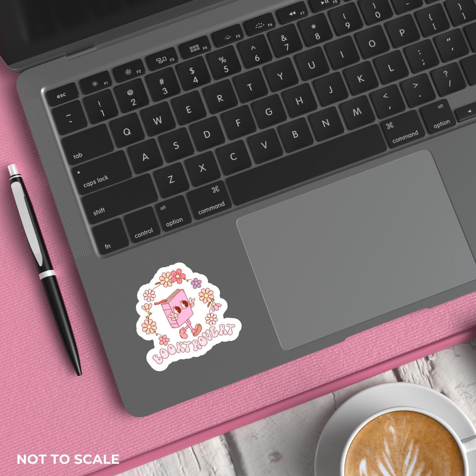 Laptop with a pink cartoon sticker on a pink surface next to a pen and coffee.