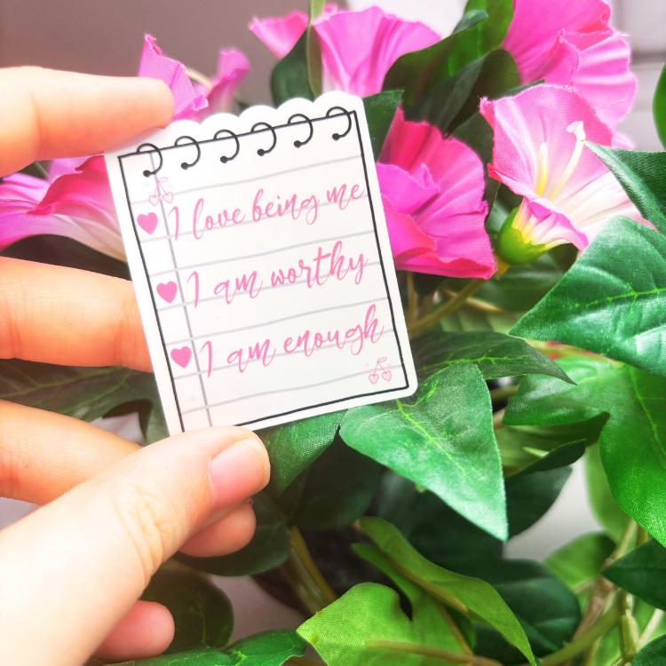 Hand holding a motivational sticker with affirmations in front of pink flowers
