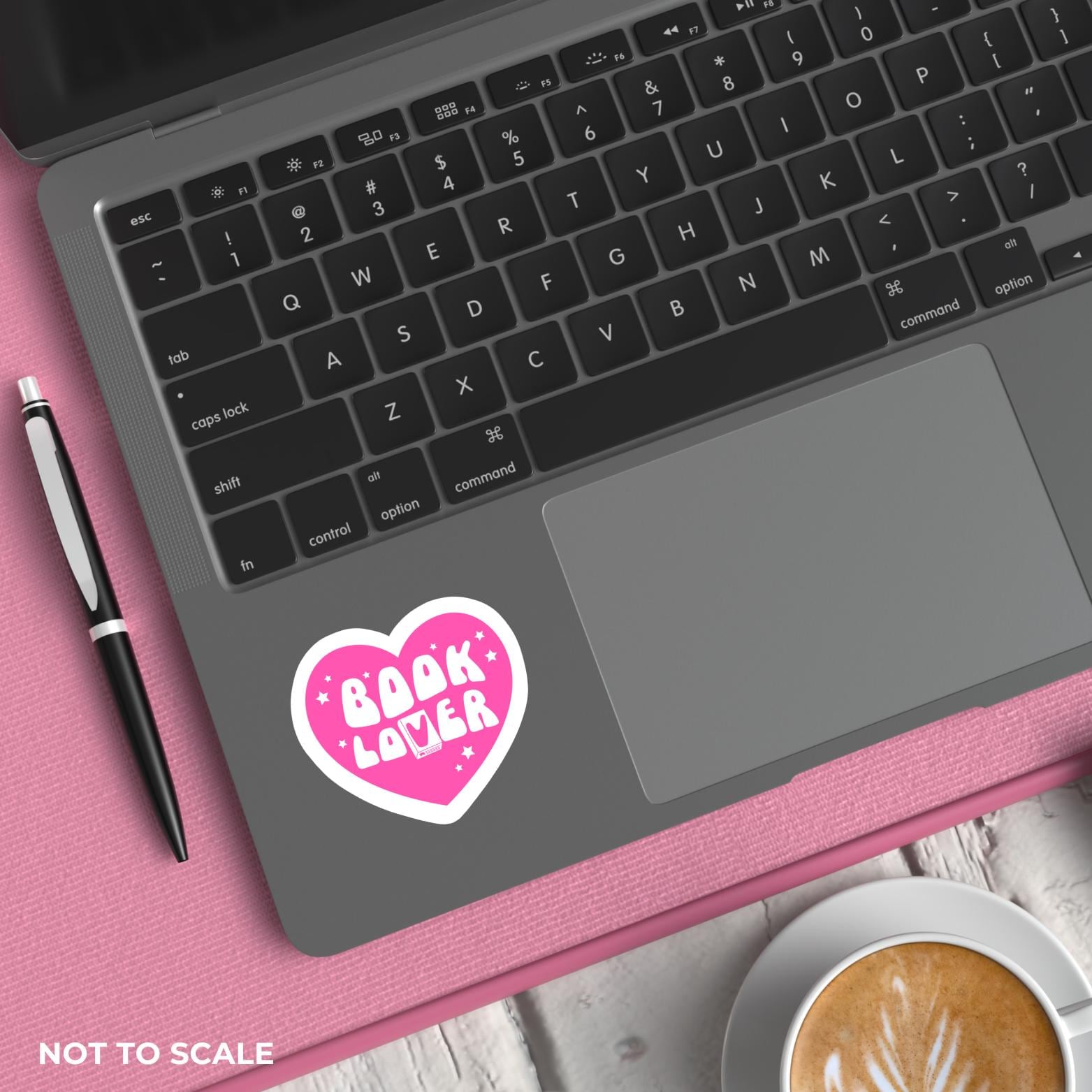 Laptop with a pink heart-shaped sticker on a pink surface with a pen and coffee cup.
