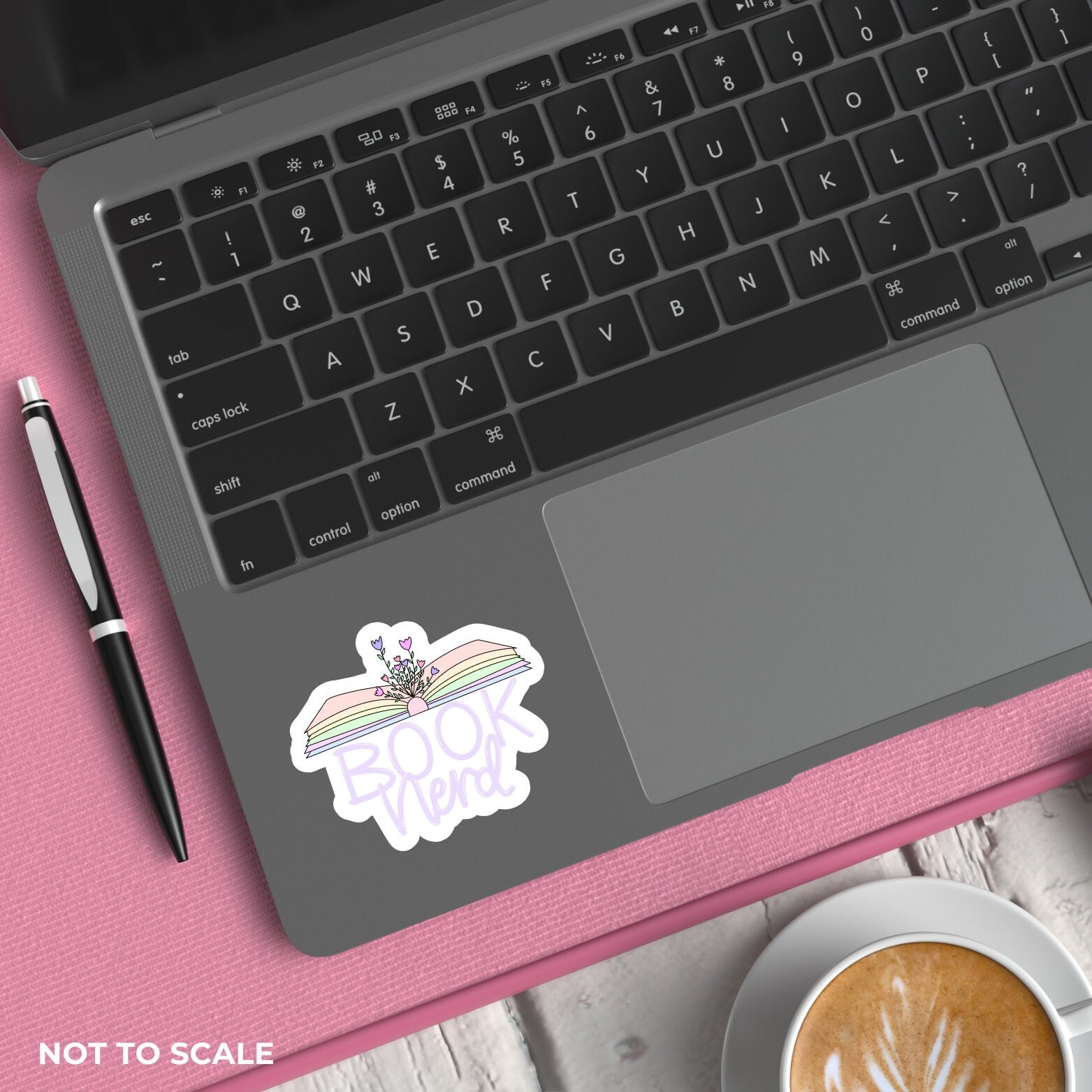 Laptop with an open book with "book nerd" text sticker on a pink surface next to a pen and coffee.