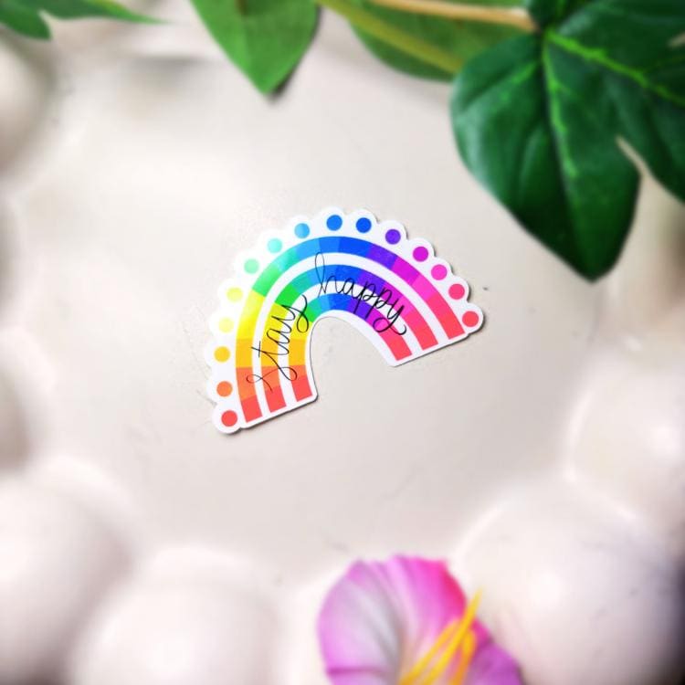 Colourful rainbow-shaped sticker with 'stay happy' text on a white surface with green leaves in the background
