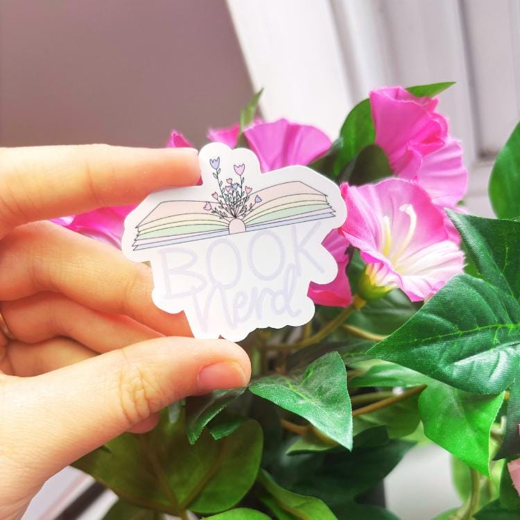 Hand holding an open book design with "book nerd" sticker with floral design in front of pink flowers