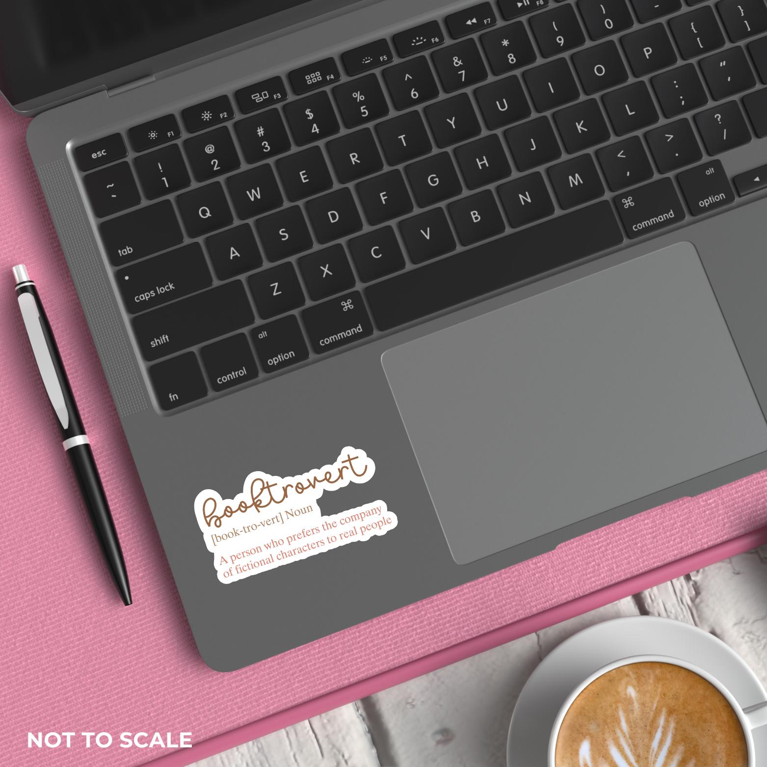 Laptop with "booktrovert" definition sticker on a pink surface next to a pen and coffee cup.