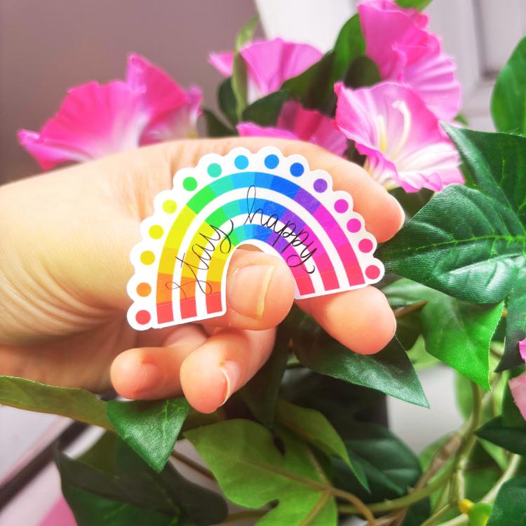 Hand holding a colourful rainbow "stay happy" sticker with pink flowers and green leaves in the background