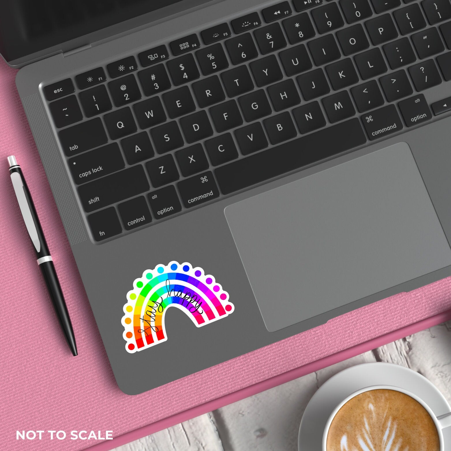 Laptop with a colourful rainbow "stay happy" sticker on a pink surface next to a pen and coffee.