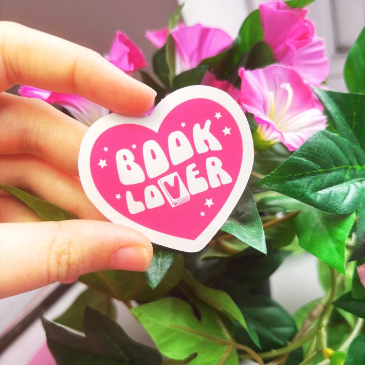 Heart-shaped 'Book Lover' sticker held by a hand with pink flowers and green leaves in the background