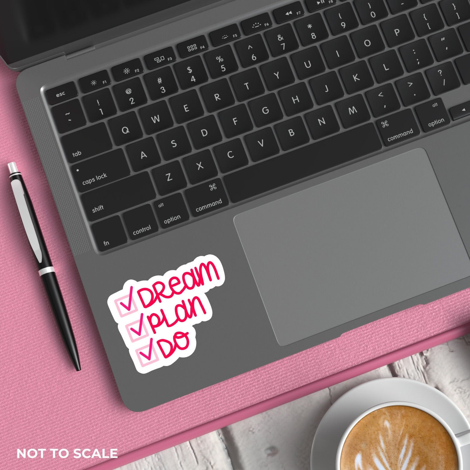 Laptop with a "dream plan do" sticker on a pink surface next to a pen and coffee.