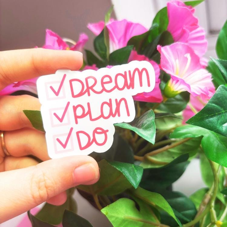 Hand holding a sticker with 'Dream, Plan, Do' text in front of pink flowers.