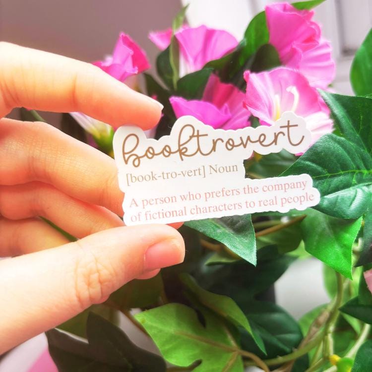 Hand holding a 'booktrovert' definition sticker with pink flowers in the background