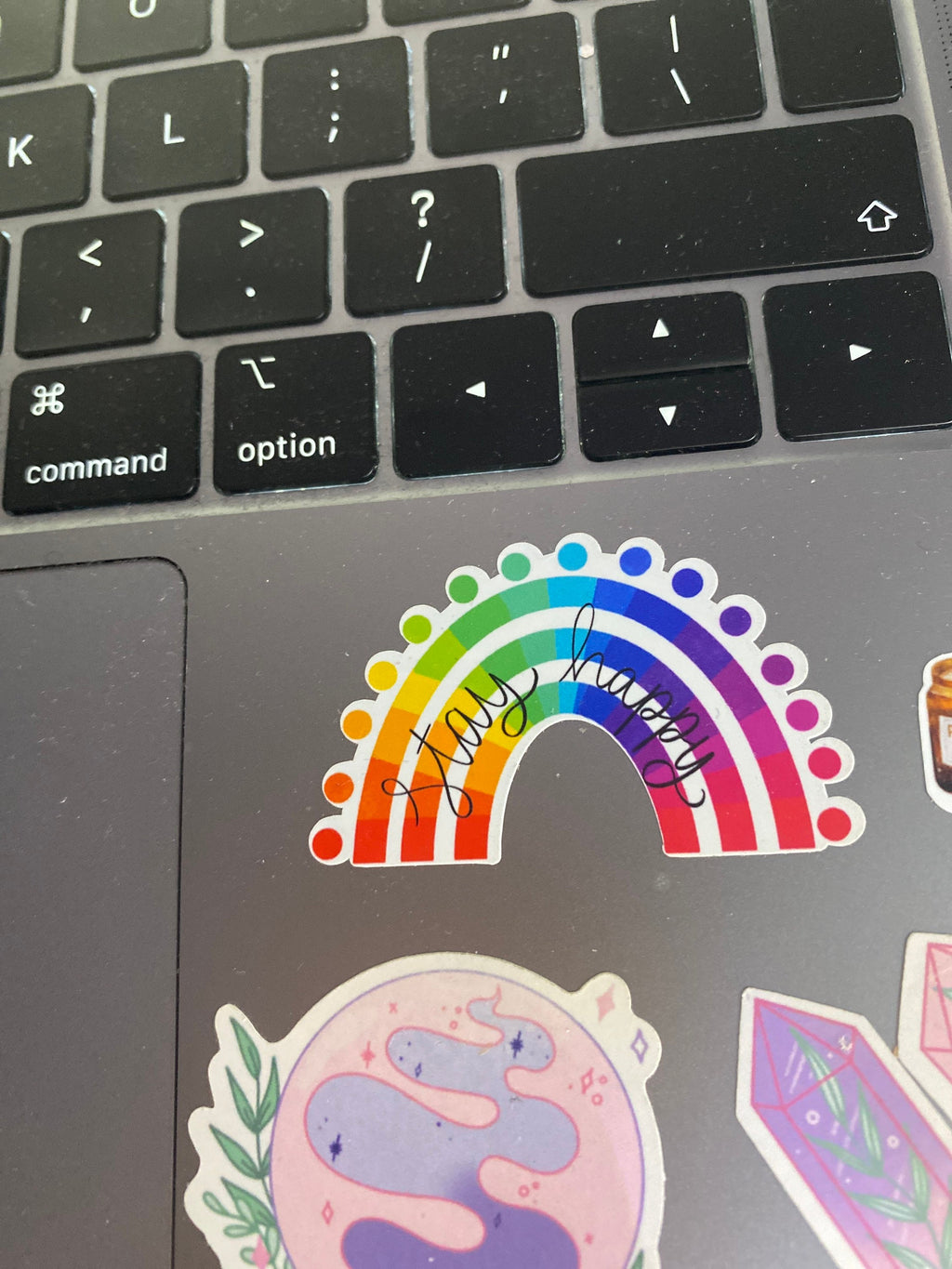 Laptop with colourful rainbow "stay happy" sticker and other decals on a black laptop palm rest.