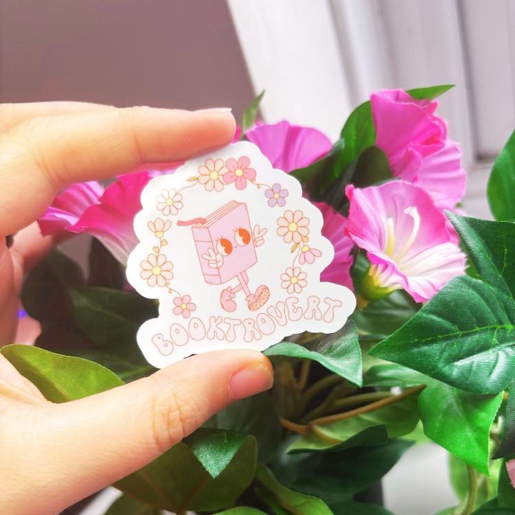 Hand holding a sticker with cartoon character and text, surrounded by pink flowers and green leaves.
