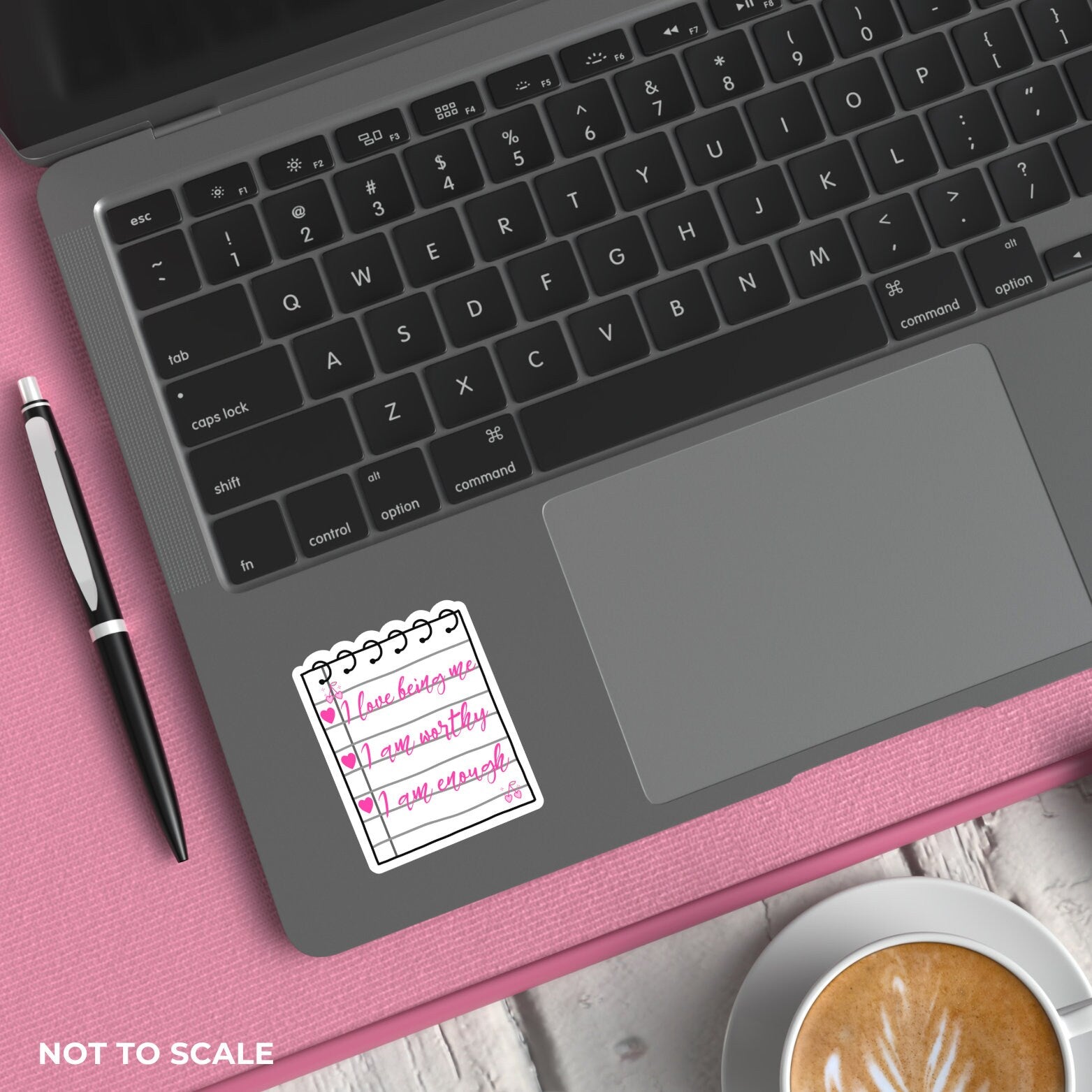 Laptop with a sticker on the palm rest area, next to a pen and coffee cup on a pink surface.